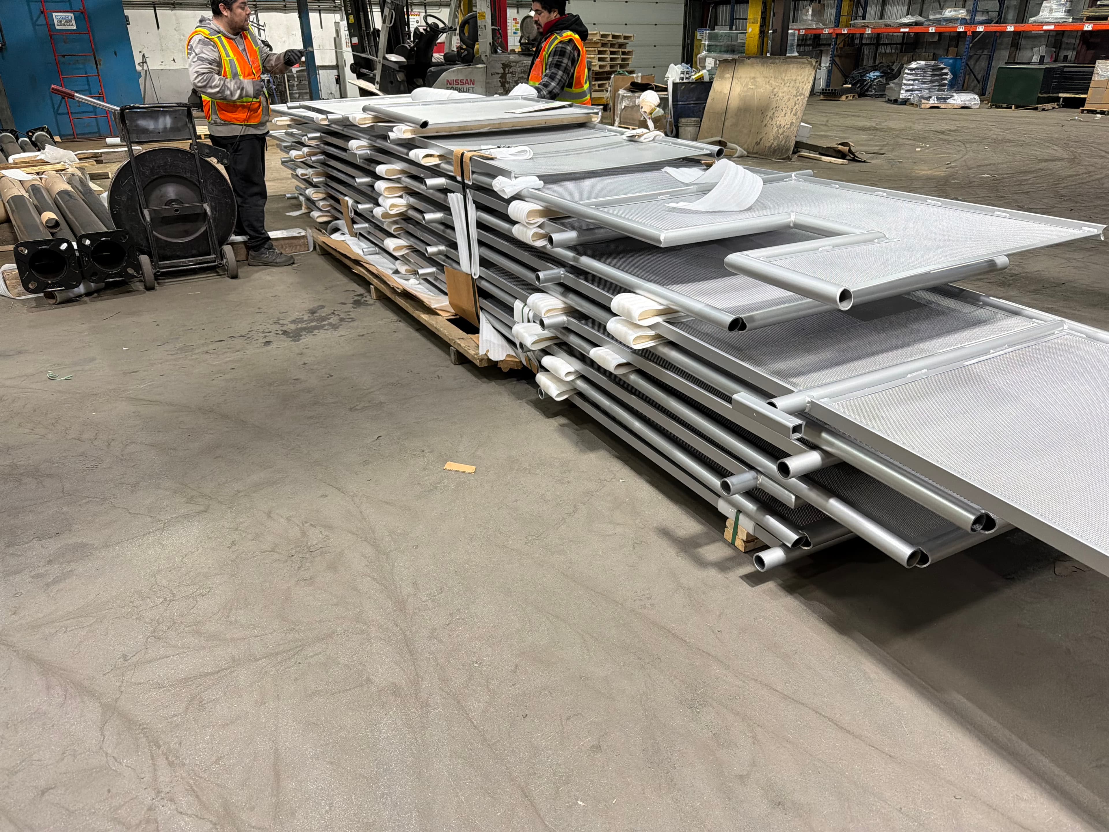 Two workers in high-visibility vests strapping a palletized stack of finished railing frames with perforated infill panels, separated by foam protection