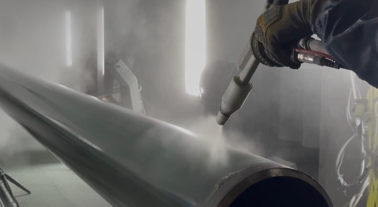 Gloved PPC coater holding a manual electrostatic powder gun, applying grey powder to the outside of a large-diameter steel pipe inside the spray booth